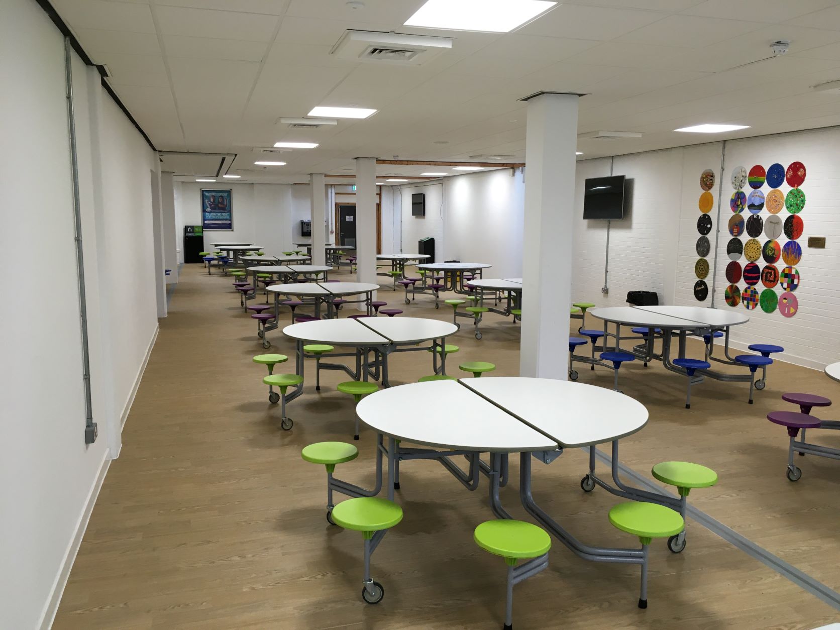 A large room with multiple circular dining tables with attached colourful round stools on wheels