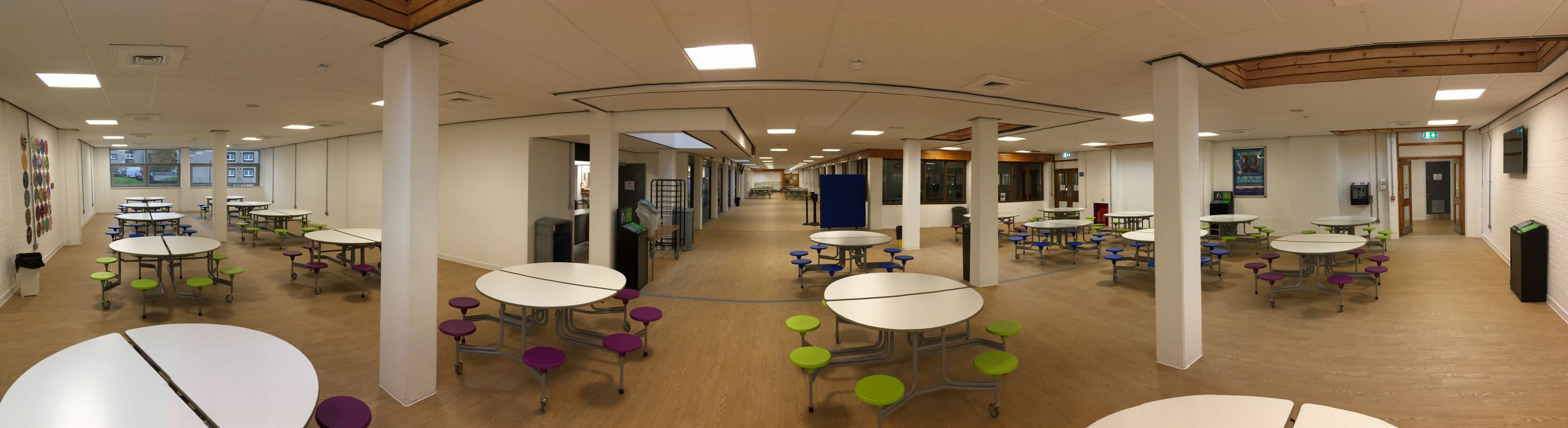 A large room with multiple circular dining tables with attached colourful round stools on wheels