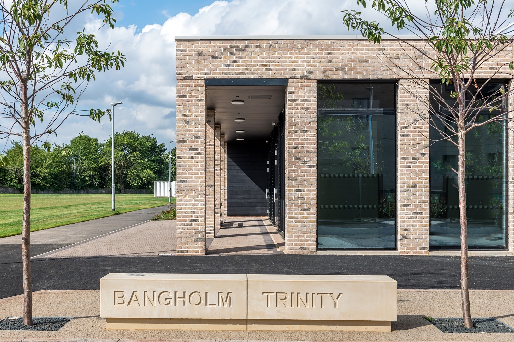 a stone bench with "Bangholm Trinity" carved into it stands in front of the brick building looking into the community room