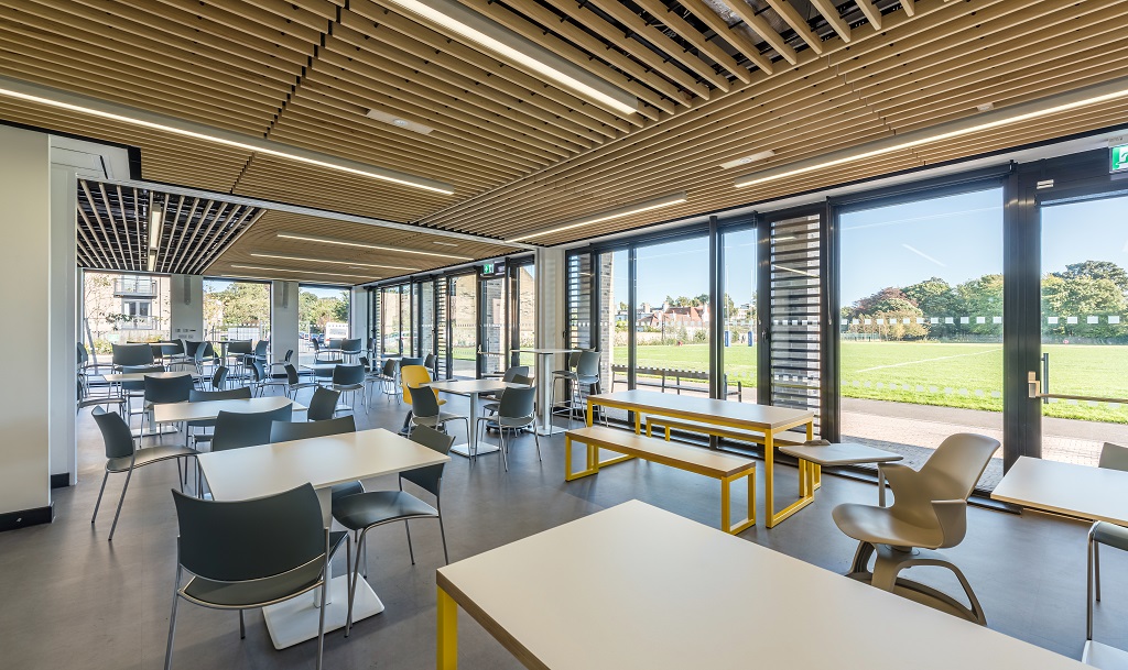 A long community room with one wall glazed looking out to the rugby pitch. Lots of different shaped tables, chairs and benching