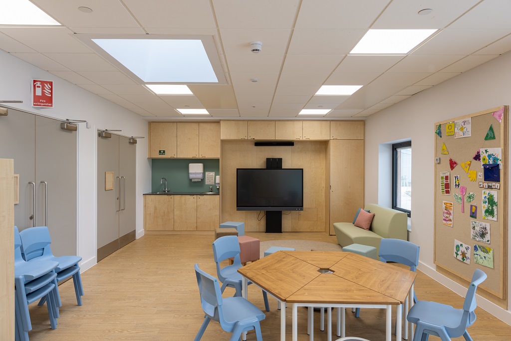 A classroom with hexagonal table and blue chairs in foreground, interactive screen on the far wall beside a sofa and stools and washing unit in background