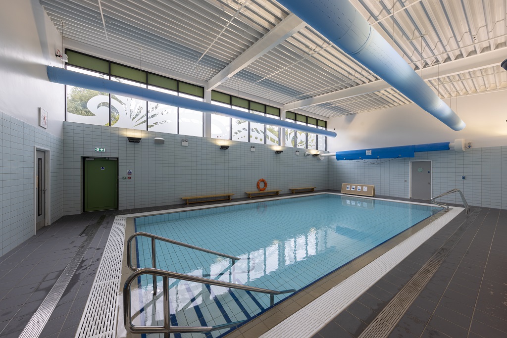 A swimming pool, in the foreground wide steps with handrails lead into the pool, the windows at the back have a leaf pattern on them