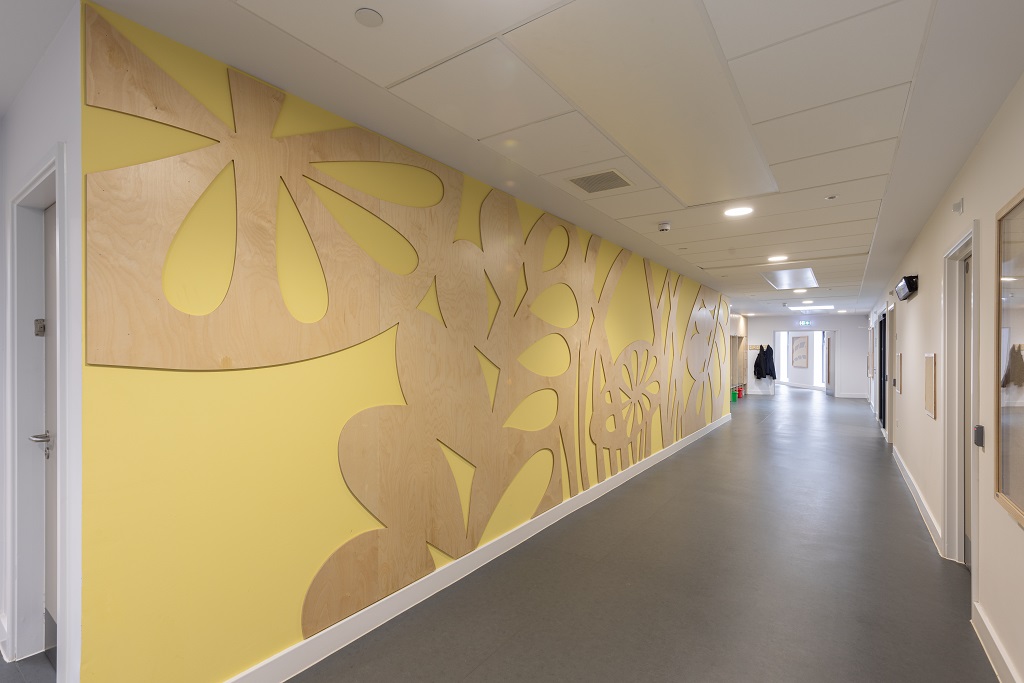 Corridor with grey flooring, the left wall is yellow with natural wood cut out relief of leaf and flower patterns