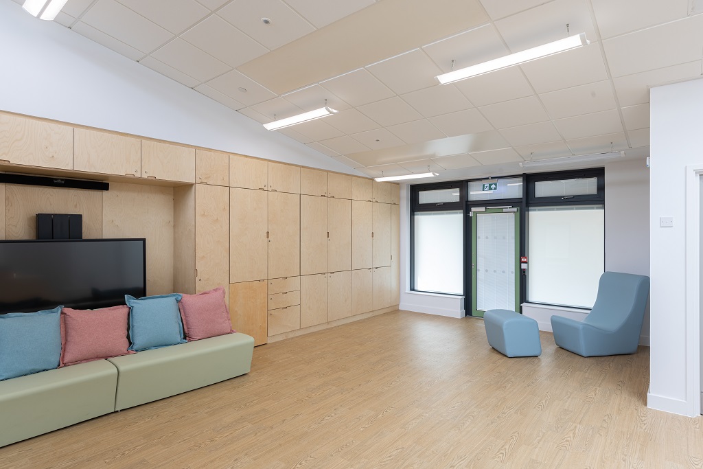 Classroom with large open floor space, bright large windows and natural wood floor and cupboards. pink and blue cushions on soft benching in front of an interactive screen and a blue armchair and footrest in the corner