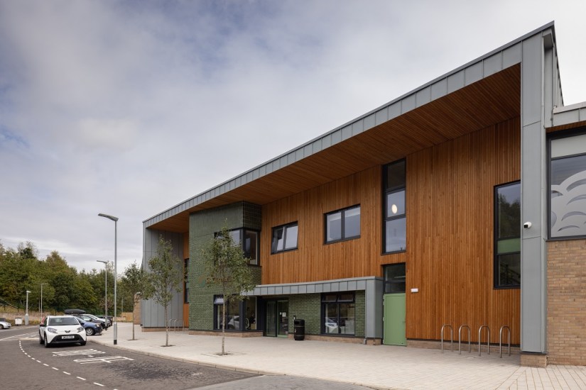 The exterior of the building's main entrance, a white car is parked in the drop off bay. The building front is mainly a warm coloured wooden cladding. A mixture of brickwork and metal gives different texture