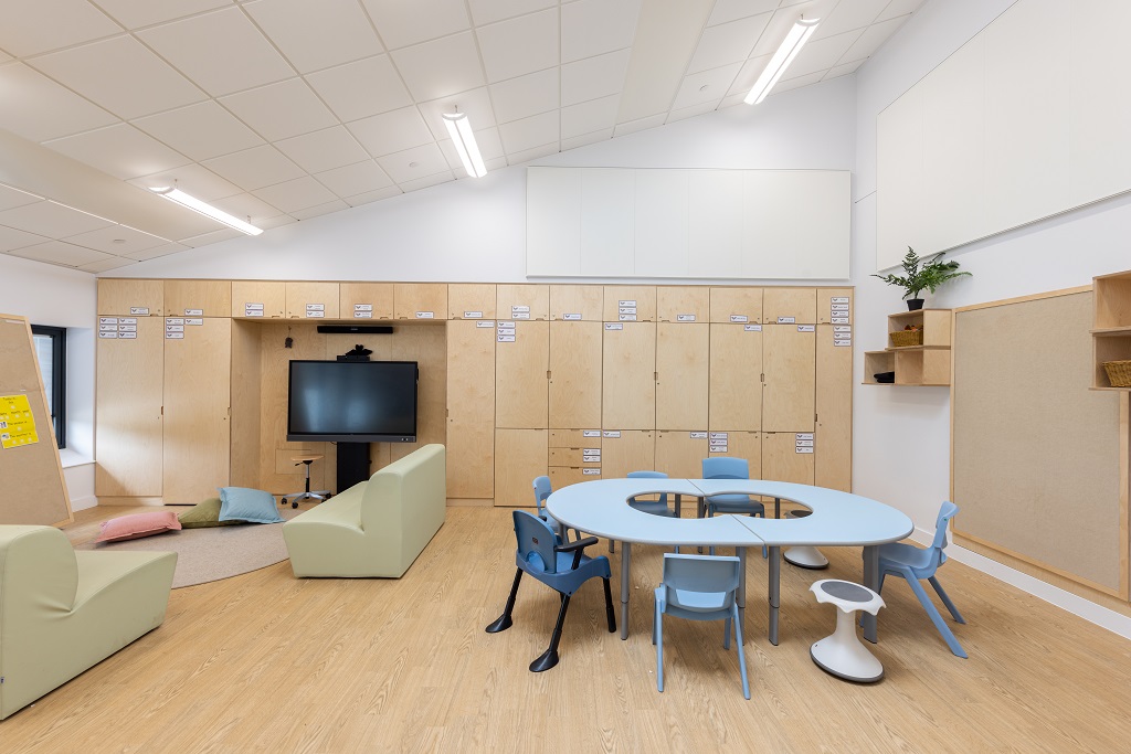 A classroom with a figure of eight shaped table and blue chairs in foreground, interactive screen on the far wall beside a couple of sofas