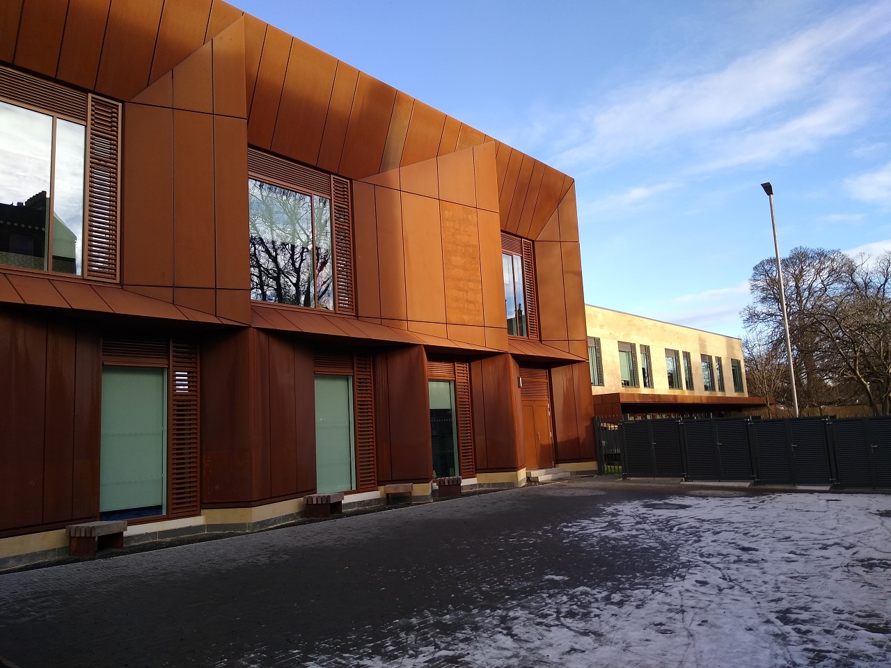The front exterior of the building, rust coloured walls with large windows, a concrete snow covered courtyard in front. Behind a low grey fence is a light stone section of the building.
