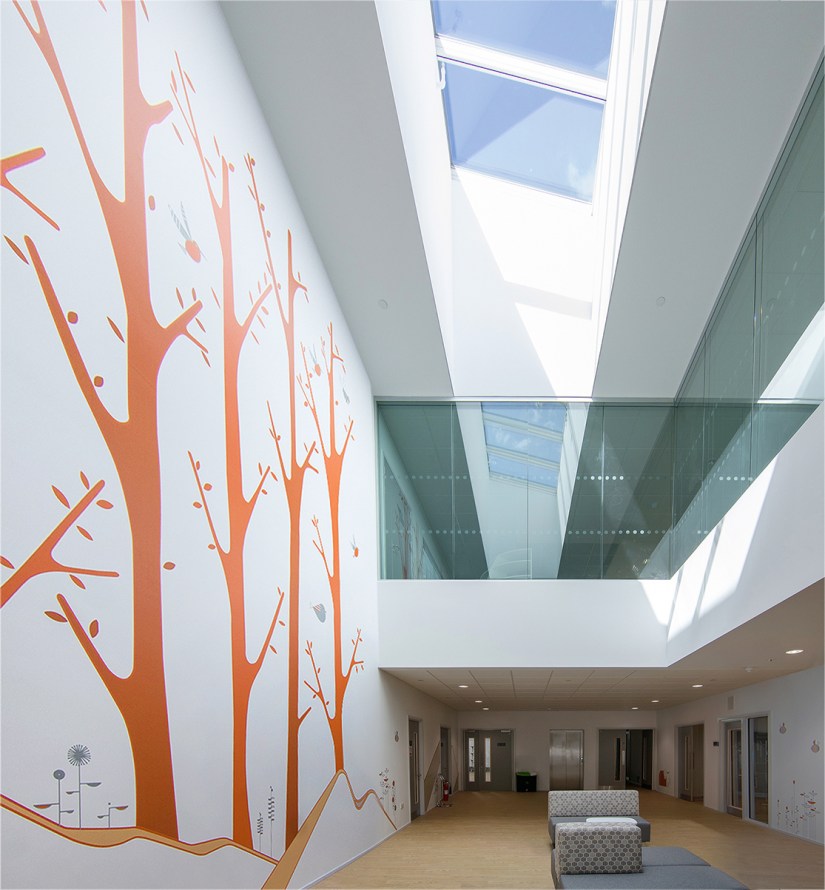 A bright two storey corridor, roof lights let in a lot of daylight. Upholstered seating is in the centre of the ground floor, on the left wall a large floor to ceiling graphic of a forest is on the wall. A glazed balcony is on the first floor.