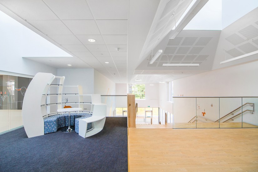 A breakout space at the top of the staircase. A rectangle of grey carpet defines the space from the natural wood style flooring of the corridor. A white U-shaped shelving unit creates a seating pod with blue soft seating within and a circular table in the centre