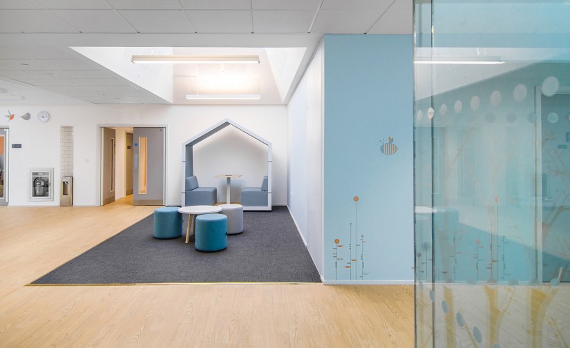 A breakout space outside the classroom, a rectangle of grey carpet defines the space from the natural wood style flooring of the corridor. Upholstered stools surround a round coffee table and a partially enclosed booth is against the wall in the background.