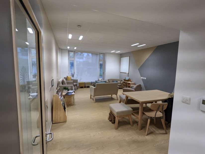 A neutrally decorated room with wooden floor and furnishings, sofas and stool by the window and a table and chairs are by the door.