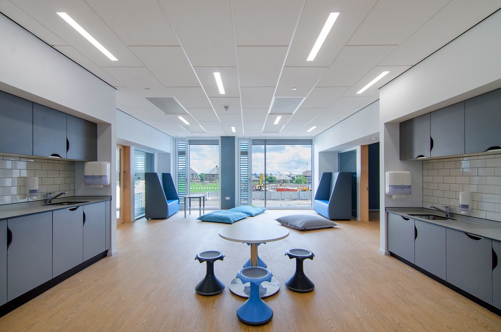 An open space between two classrooms, on either side is a worktop and cabinets and a sink. In the centre is a round table and four wobble stools. At the back of the room beside a glazed wall are two high backed sofas and some large floor cushions