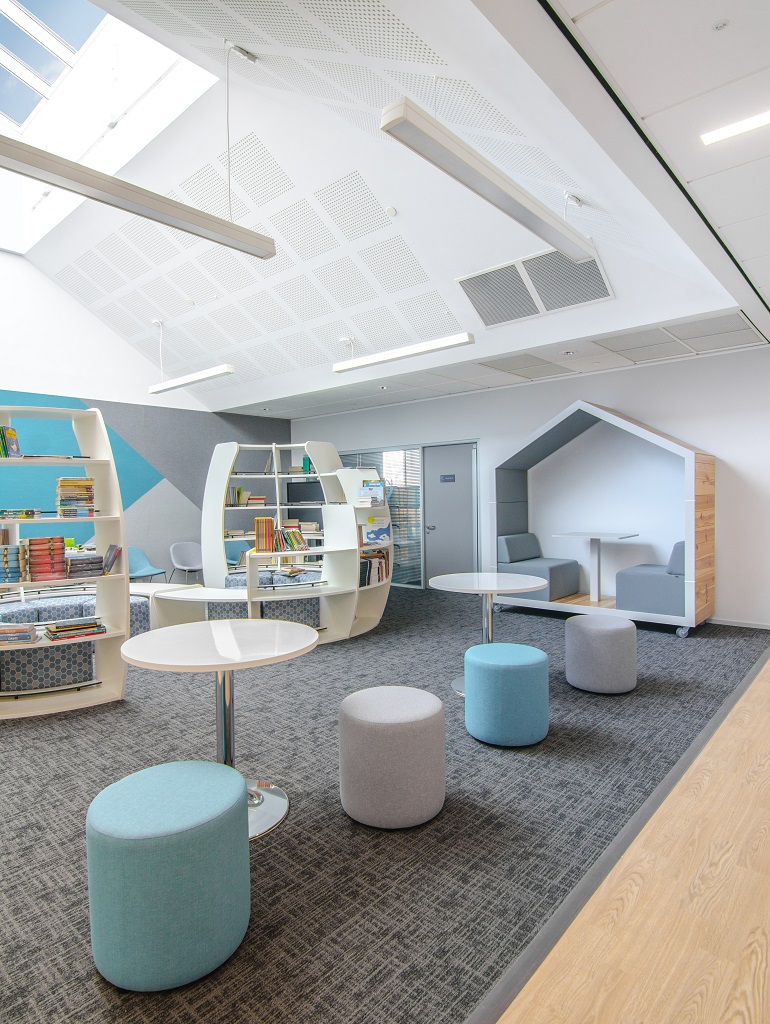 A light and bright open space. Grey patterned carpet marks out the library space, a couple of round tables and upholstered stools in the foreground, behind curved bookshelves and a partially enclosed booth