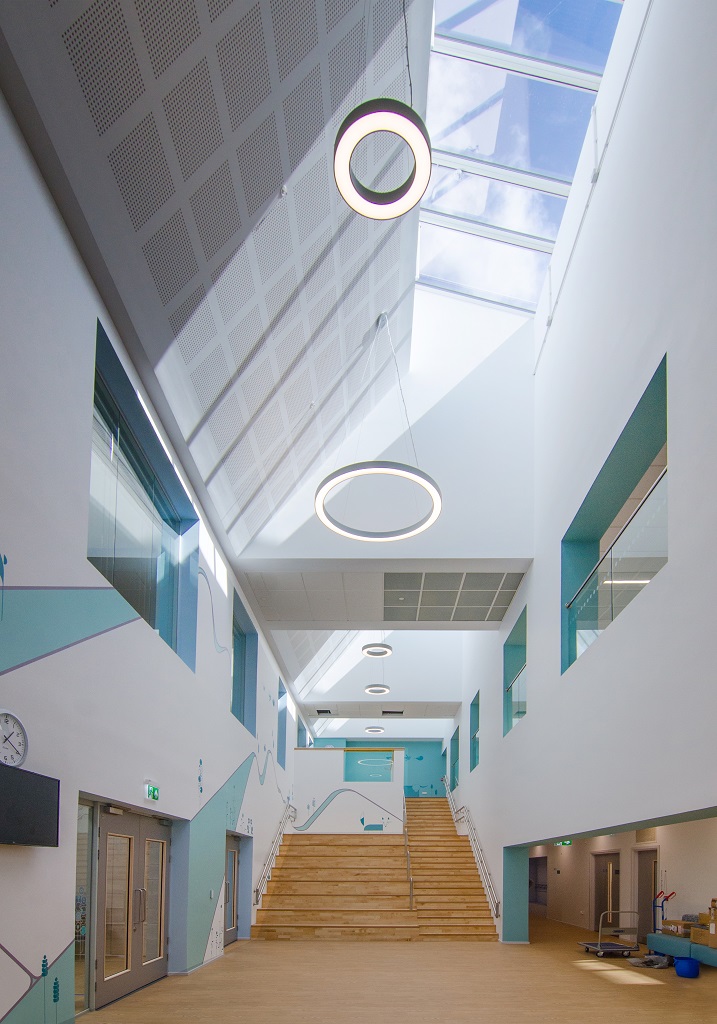 The school foyer, a two storey space with roof lights creating a light and bright room. A staircase and hellerup stair lead to the first floor with a balcony on the right and windows on the left. A blue illustration is on one wall.