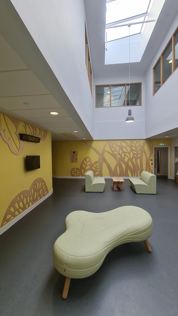 An open space between rooms, in the foreground is a rounded triangular stool, in the background two sofas facing eachother with a coffee table between. The room has grey flooring, the walls are yellow with natural wood cut out relief of leaf and flower patterns