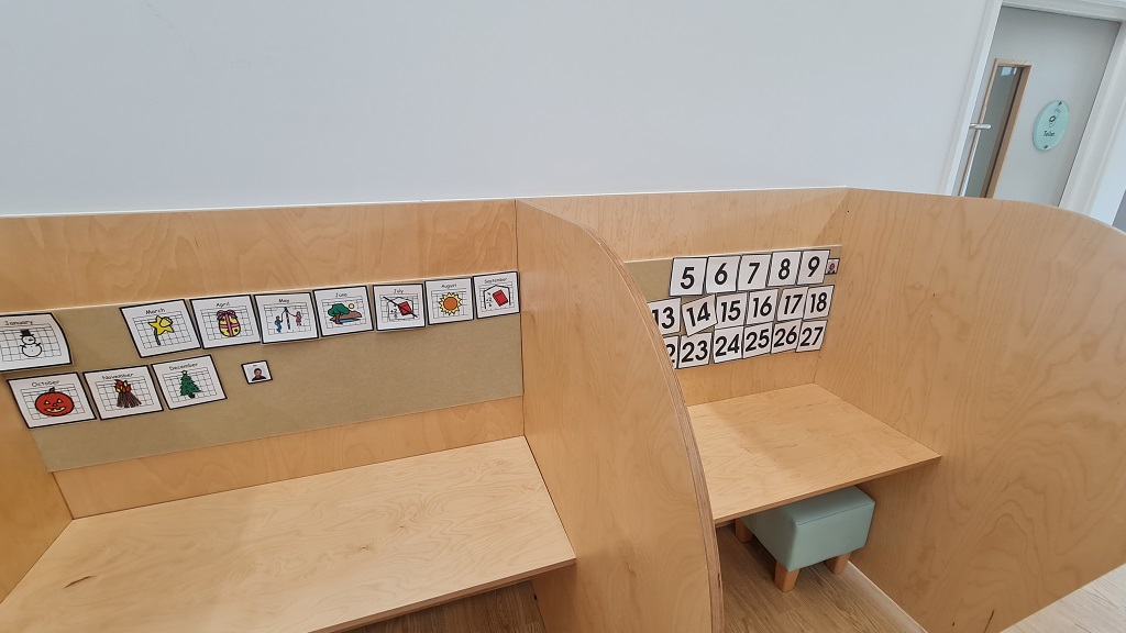 Two natural wood focus booths, a square stool sits under a desk with three high sides. Numbers and images are stuck onto the pinboarding on the central wall of the booth