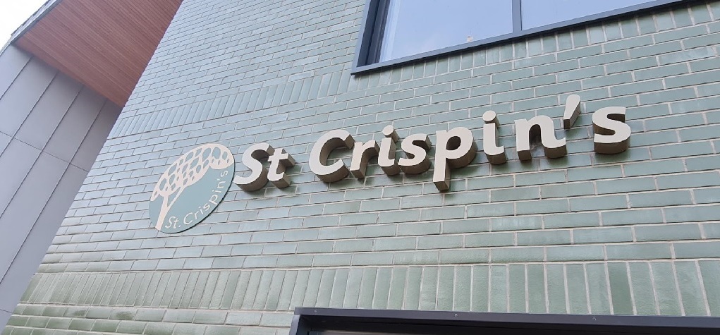 A brick wall with a circular metal sign showing a tree and "st crispin's" beside a metal embossed lettering of the name of the school