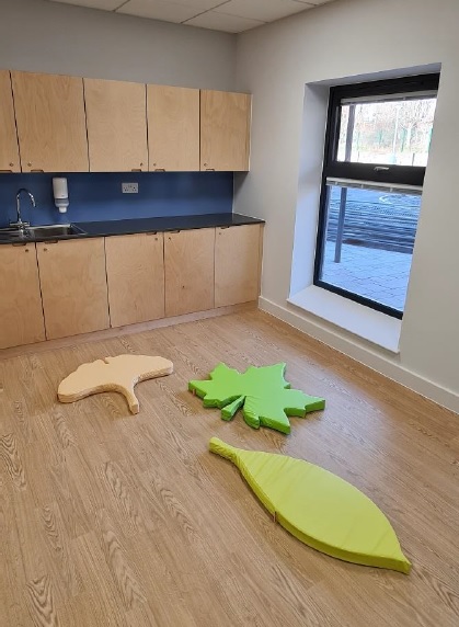 A playroom with natural wood floor and storage cupboards with sink, colourful leaf shaped soft cushions are on the floor