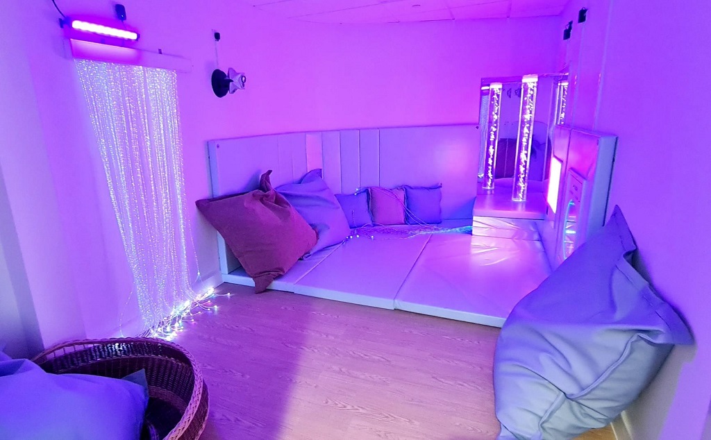 A room lit purple, a mixture of large and small cushions are against the walls, a basket, cushioned floor and wall panels and LED strings and bubble lamp