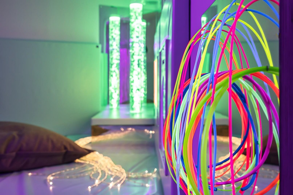 A close up of rainbow coloured tubes, a cushion, lit LED strings and bubble lamp lit up in front of a mirror