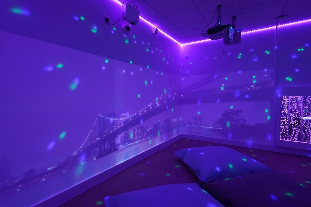 A room in dark purple light, with projectors displaying images on the walls of a city scape with bridge at night, a light panel is on the right side wall