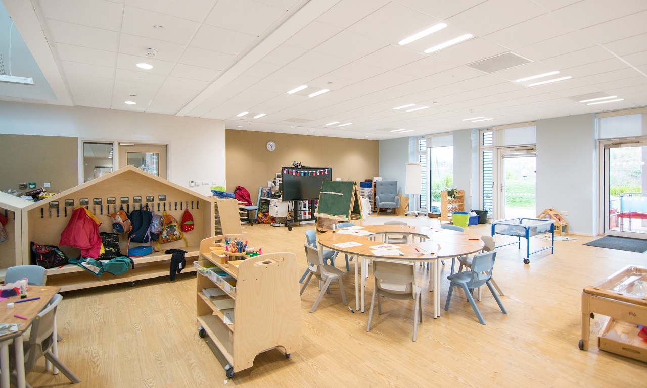 A neutrally decorated classroom, house shaped coat racks, circular tables and wooden activity furnishings and storage