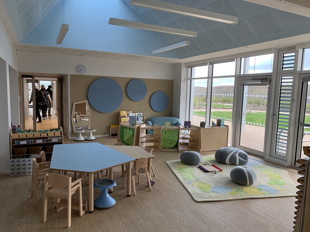 A classroom with light wood flooring and furnishings, bean bags that look like pebbles on a rug, blue circles on the wall and a blue pentagon table with chairs around, floor to ceiling windows