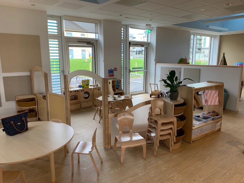The nursery home corner with natural wood furniture, children's chairs are stacked, low dividers partition the room into activity areas