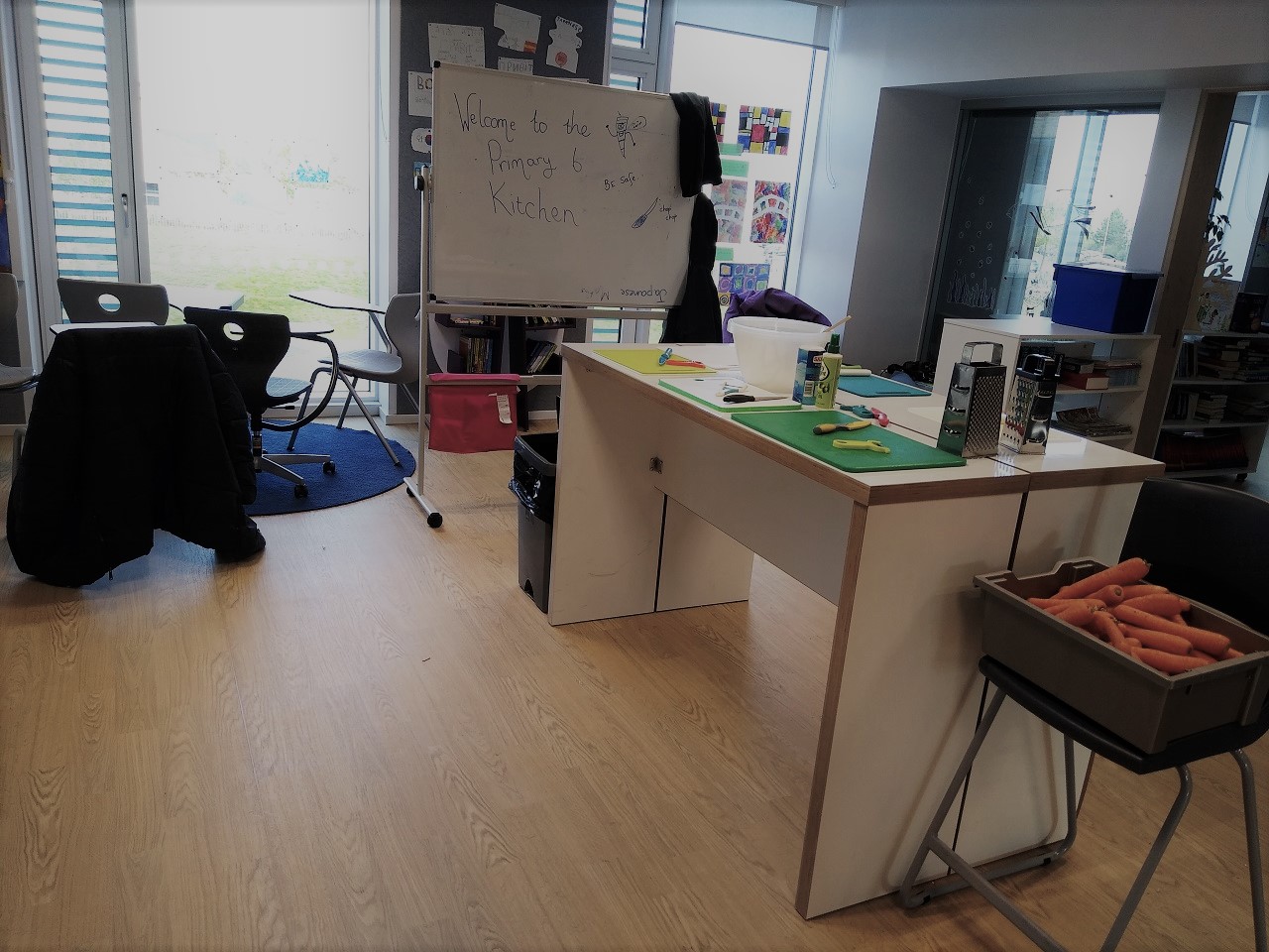 A breakout space, a white board says "welcome to the Primary 6 kitchen" in front is a white standing desk laid out with chopping boards, peelers, graters and a box of carrots is on a stool.