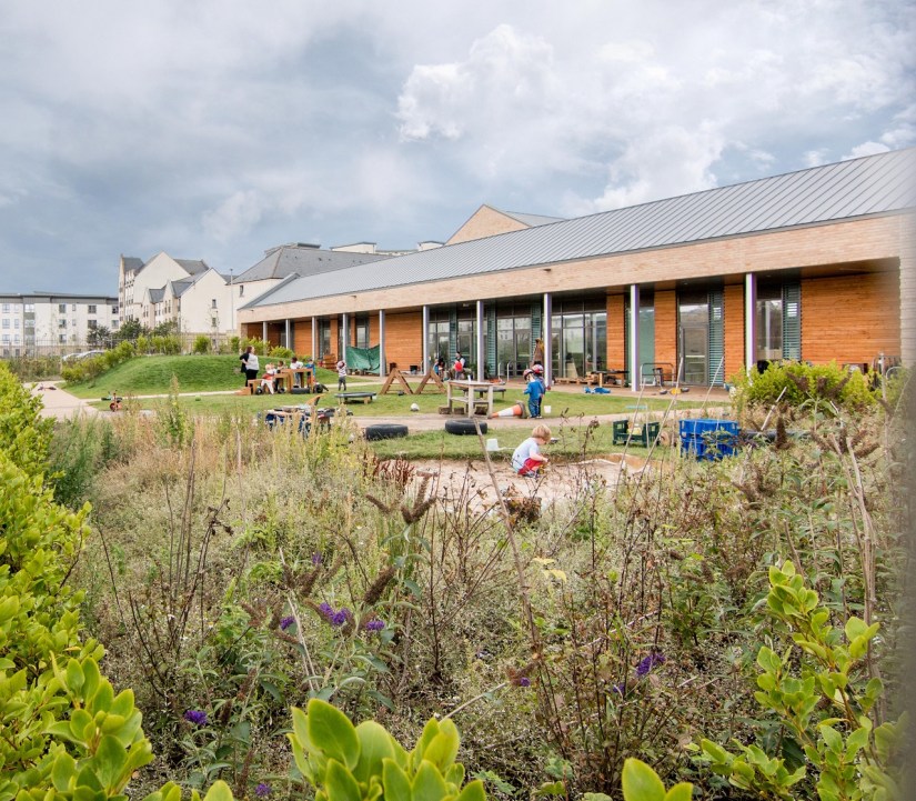 A green playground with children playing, a sandpit, paths and small hill in front of the nursery building